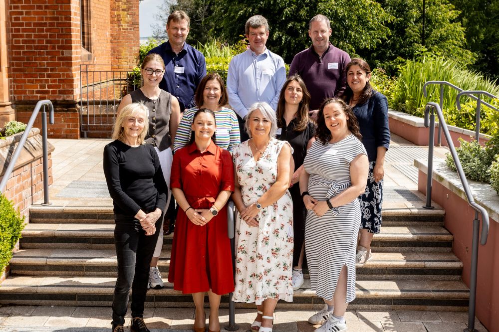 Eleven conference attendees, three male and eight female, standing in three rows on some steps outside a red brick building in the sunshine.